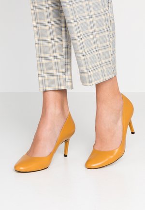 Gele leren hoge hakken met een gladde afwerking en een puntige neus, geplaatst tegen een lichte structuur van een grijze geruite broek.