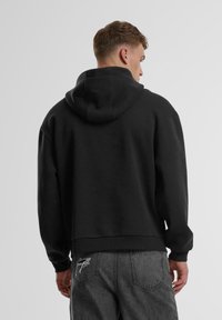 Schwarzer Kapuzenpullover mit lockerer Passform, aus strukturiertem Stoff und mit gerippten Bündchen. Die Rückansicht zeigt eine verkürzte Länge und keine sichtbaren Logos.