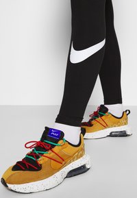 Nike-sneakers med en kombination av gul mockaband och svarta accenter, med multikolorade snören och en spräcklig vit sula, visade tillsammans med svarta leggings.