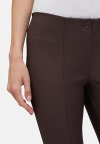 Brązowe spodnie z rozciągliwej tkaniny o gładkiej fakturze, z przodem zapinanym na guzik i szwami bocznymi, pokazane z ręką opartą na talii.