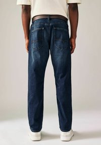 Dark blue denim jeans with a straight-leg cut, visible back pockets, and contrasting stitching. Worn with a white belt and sneakers.