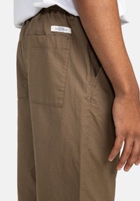 Brown pants with a textured fabric, elastic waistband, and a back pocket with a label. The design features a loose fit and simple lines.
