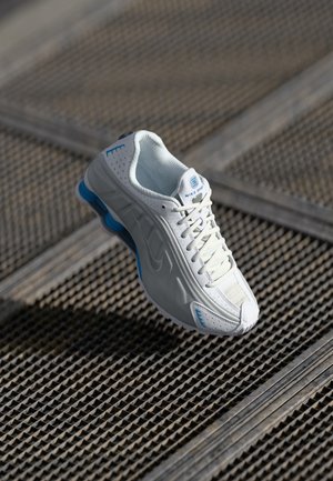 Hvid og blå Nike løbesko med snørebånd, svævende over en tekstureret metalristeflade i naturligt lys.