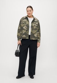 Veste florale camouflage en verts avec deux poches poitrine, associée à un pantalon large noir et un petit sac noir.