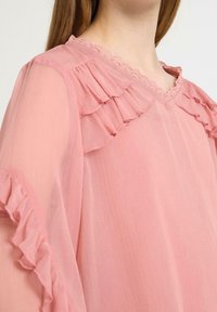 Rosa Chiffonbluse mit Rüschenakzenten entlang des Ausschnitts und der Ärmel; verfügt über einen V-förmigen Spitzenbesatz für zusätzliche Details und Textur.