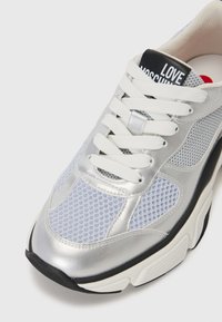 Srebrni in svetlo modri športni copati z mrežastimi paneli, belimi vezalkami in črnimi etiketami z napisom "LOVE MOSCHINO". Debel podplat.