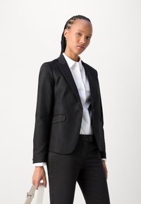 Černý blazer s hladkou texturou a upraveným střihem, spárovaný s bílou košilí s límečkem. Modelka drží béžovou tašku.