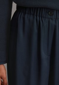 Marineblauwe broek met elastische tailleband en knoopsluiting. De stof lijkt glad met een subtiele glans. Bevat zijzakken en heeft een rechte snit.