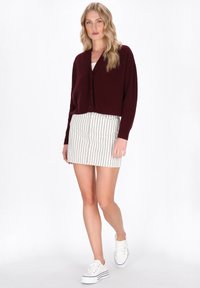Bordeaux neulecardigan, jossa on V-pääntie ja nappisuljenta, paritettu valkoisen raidallisen mini-hameen ja valkoisten tennareiden kanssa.