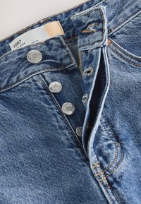 Primer plano de jeans de mezclilla azul con cuatro botones metálicos visibles y costuras marrones alrededor de los ojales y las costuras.
