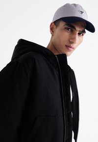 Chaqueta negra con capucha y cremallera, con un gran bolsillo frontal. Gorra gris y azul marino con logotipo bordado. Textura de tela suave.