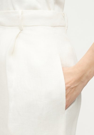 Χέρι που ακουμπάει μέσα στην πλαϊνή τσέπη λευκών παντελονιών από λινό με ψηλή μέση και θηλιά για ζώνη.