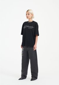 Camiseta negra de gran tamaño con texto bordado en plata, combinada con unos pantalones grises holgados adornados con coloridos bordados de flores y zapatos negros de punta.