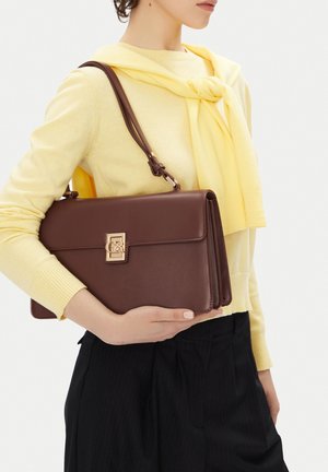 Žena v žlutém svetru drží tmavě hnědou koženou kabelku přes rameno se zlatou sponou, oblečená v černých kalhotách, stojí proti bílému pozadí.