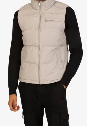 Homme portant une veste matelassée beige clair par-dessus un pull noir à manches longues et un pantalon cargo noir, une main dans la poche.