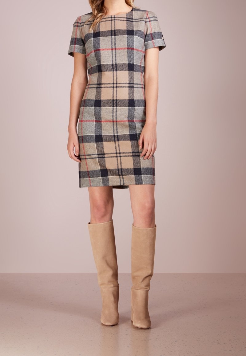 Ruitenkleurige jurk met korte mouwen in beige, grijs en zwart met rode accenten. Knielange snit gecombineerd met beige suède laarzen tot de knie.