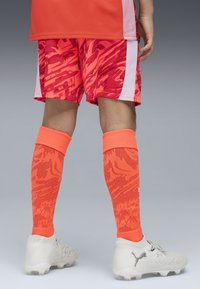 Tenue de sport composée d'un t-shirt orange vif, de shorts rouges à motifs et de chaussettes longues orange assorties. Les chaussures sont des crampons de football gris.