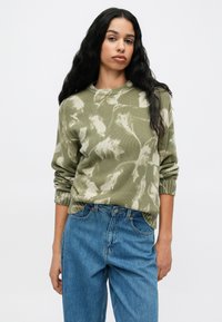 Kaki žalios spalvos raštuotas megztinis su apvaliu iškirpimu ir raukšlėmis rankogaliuose, derinamas su šviesiai mėlynomis džinsinėmis kelnėmis. Tekstūruotas, laisvas kirpimas.