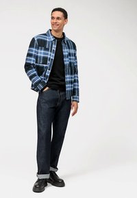 Giacca a quadri blu con maniche nere, indossata sopra una camicia nera. Jeans in denim scuro risvoltati alle caviglie, abbinati a scarpe nere.