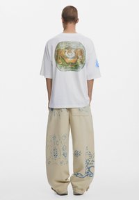 Laisvas baltas marškinėlis su ryškiu tigro grafikos piešiniu nugarinėje pusėje, derinamas su smėlio spalvos kelnėmis, papuoštomis mėlynais raštais ir akcentais.