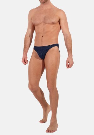 Marineblaue Schwimmhose mit weißen Punkten, aus glattem Stoff gefertigt. Verfügt über einen tiefen Schnitt und seitliche Öffnungen für minimale Bedeckung.