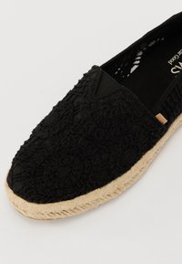 Sorte espadrille med teksturert stoff som har blomstermønstre. Designet inkluderer en tau-såle og en myk, flat form.