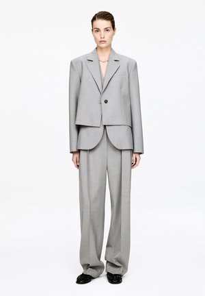 Kvinde stående iført oversize lys grå jakkesæt med enkeltknappet blazer og vidde bukser, kombineret med sorte sko og en kort halskæde.