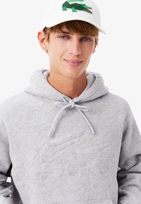 Felpa grigia con texture floreale, cordino regolabile e tasca a marsupio. Indossata con un cappellino bianco con un logo di coccodrillo verde.