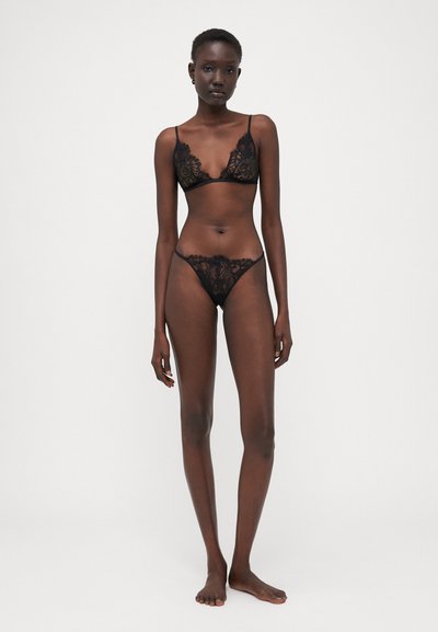 Conjunto de lencería de encaje negro que incluye un bralette con delicados bordes en forma de onda y un thong a juego, ambos con patrones florales transparentes.