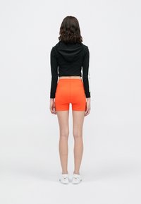 Sweatshirt cropped preto com mangas longas e riscas laterais brancas; calções justos laranja brilhante; sapatilhas brancas.