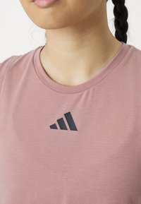 Lyserød atletikshirt med rund halsudskæring og korte ærmer, prydet med et sort geometrisk logo på brystet. Blød tekstur, afslappet design.