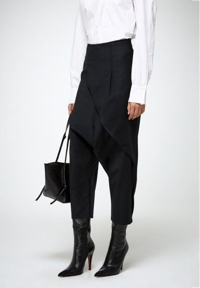 Zwarte hooggetailleerde broek met asymmetrische draping, gecombineerd met elegante zwarte stiletto laarzen en een kleine zwarte handtas.