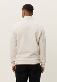 Μπεζ φούτερ με ψηλό γιακά και ριμπ μανσέτες, με λεία υφή και εφαρμοστή σιλουέτα. Χωρίς εμφανή λογότυπα ή σχέδια.