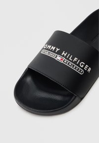 Fekete csúszásmentes szandál, széles pánttal, rajta a "TOMMY HILFIGER" logó fehér és piros színben. Texturált talp, sima pánt anyag, minimális design.