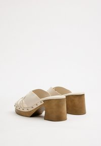Zapatos de tacón plataforma beige con tiras cruzadas, acentos de tachuelas doradas y un tacón de bloque de madera marrón de forma única. Textura suave.