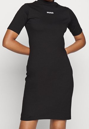 Černé krátké přiléhavé šaty s krátkým rukávem a vysokým výstřihem, s bílým logem "HUGO" uprostřed na hrudi, nošené osobou s rukama za zády.