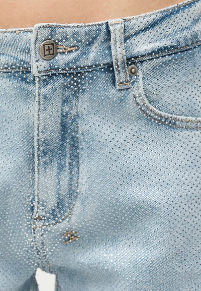 Ανοιχτό μπλε τζιν με μικρές ασημένιες τρουκς που καλύπτουν το ύφασμα, με κουμπί και κούμπωμα κοντά στην μέση και στην μπροστινή τσέπη.