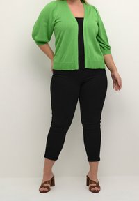 Femme portant un cardigan en tricot vert vif, un haut noir, un pantalon slim noir et des sandales à talons ouverts marron, se tenant devant un fond uni.