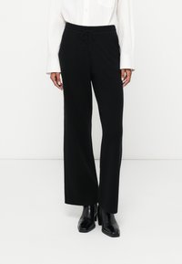 Černé kalhoty s širokými nohavicemi vyrobené z měkk�é tkaniny s páskem na stažení v pase, v kombinaci s bílou košilí na knoflíky a černými botami na podpatku.