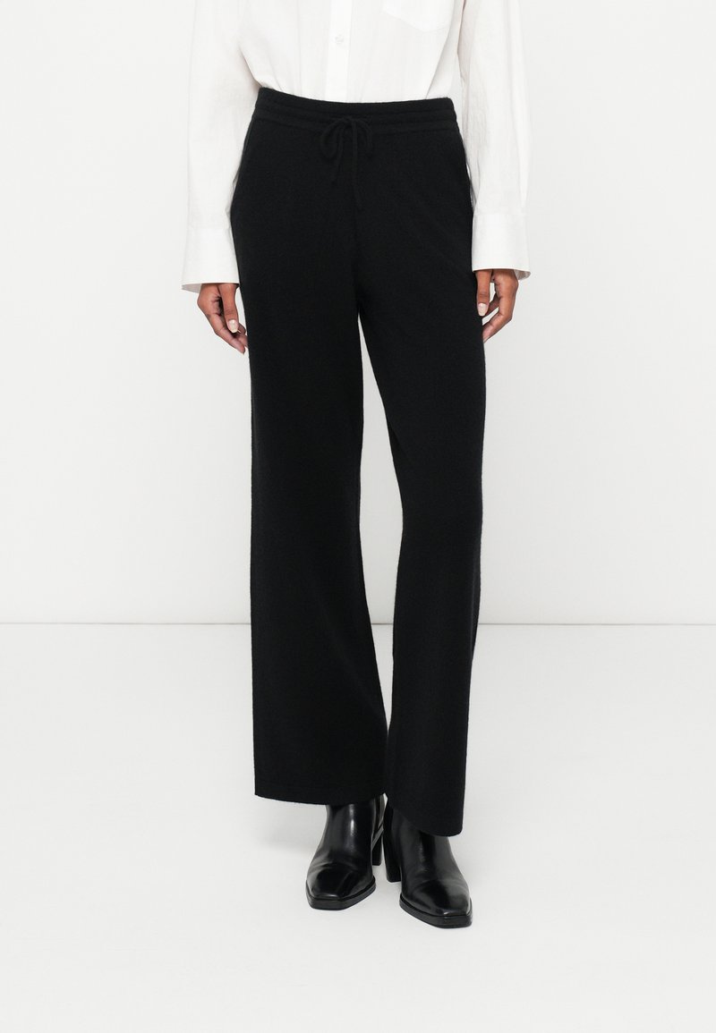 Zwarte wijde broek gemaakt van een zachte stof met een trekkoord in de taille, gecombineerd met een witte overhemd met knopen en zwarte laarzen met hak.