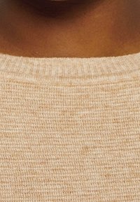 Pull en tricot beige avec un col rond côtelé, présentant un tissu texturé et léger ainsi que des variations subtiles de couleur.