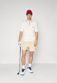 Ung mann i golfantrekk med rød caps, hvit polo, beige shorts, hvite hansker og holder en golfkølle, står på ensfarget bakgrunn.