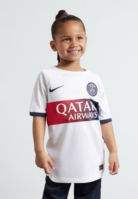 Camiseta de fútbol blanca con acentos en azul y rojo; presenta el logo de "QATAR AIRWAYS" y el logo de París. Mano de un niño reposando en la cadera, sonriendo.