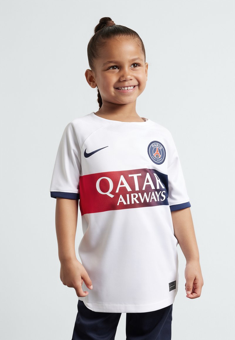 Camiseta de fútbol blanca con acentos en azul y rojo; presenta el logo de "QATAR AIRWAYS" y el logo de París. Mano de un niño reposando en la cadera, sonriendo.