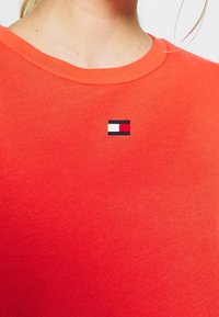 Gros plan d'une personne portant un t-shirt côtelé orange à col rond avec un petit logo rectangulaire rouge, blanc et noir centré près du col.