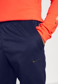 Nike dukserice u tamnoplavoj boji s elastičnom trakom u struku i bočnim džepovima. Na lijevoj nozi ima crni logo. U paru s dugim rukavom u jarkoj narančastoj boji.