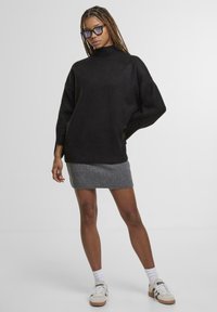 Černý oversize svetr s rolákem a dlouhými rukávy, v kombinaci se šedou texturovanou mini sukní. Modelka má na sobě bílé boty a brýle.