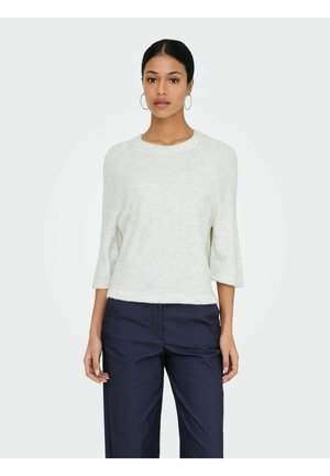 Femme aux cheveux foncés portant un haut blanc à manches longues et un pantalon bleu foncé, debout devant un arrière-plan clair uni, faisant face à l'avant.