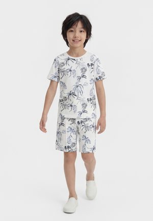 Dziecko idzie do przodu, nosząc biały dopasowany t-shirt i szorty z niebieskimi i czarnymi nadrukami palm oraz białe wsuwane buty.