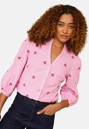 Woman with curly hair wearing a pink button-up blouse with red embroidered bows and dark high-waisted pants, hand in pocket, neutral background.